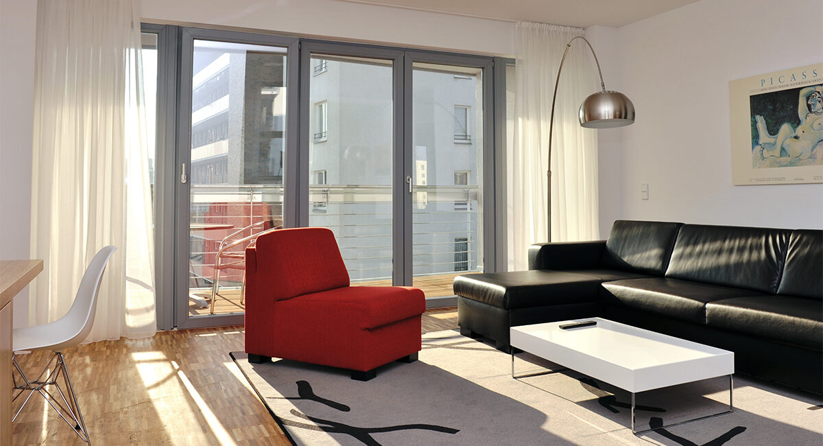 Ein geräumiges, sonniges Wohnzimmer in einer Wohnung im Grimm's Hotel in Berlin Mitte mit einem schwarzen Sofa, einem roten Sessel und großen Fenstern.